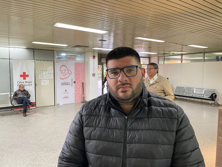 Hombre de barba con chaqueta negra y gafas, esperando en un espacio público con carteles de la Cruz Roja en las paredes.