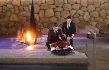 Rubén Ramírez Lezcano y Pedro Alliana en traje, colocando ofrenda circular con flores en ambiente ceremonial con fogata al fondo.