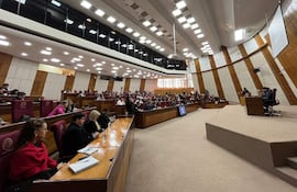 Asistentes formales en audiencia pública, algunos con expresión tranquila y otros tristes, mientras un orador habla en el estrado.