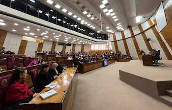 Asistentes formales en audiencia pública, algunos con expresión tranquila y otros tristes, mientras un orador habla en el estrado.