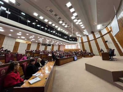 Asistentes formales en audiencia pública, algunos con expresión tranquila y otros tristes, mientras un orador habla en el estrado.