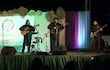 Dos guitarristas y un bajista en chaquetas oscuras tocan en un escenario decorado con telas coloridas durante el festival.
