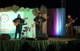 Dos guitarristas y un bajista en chaquetas oscuras tocan en un escenario decorado con telas coloridas durante el festival.