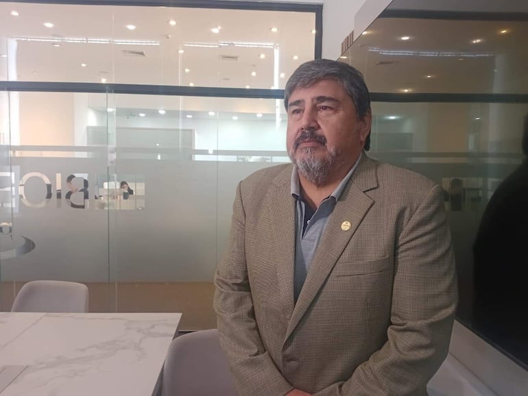 Villalba de pie en saco sobre camisa gris, con fondo de personas trabajando en oficina moderna.