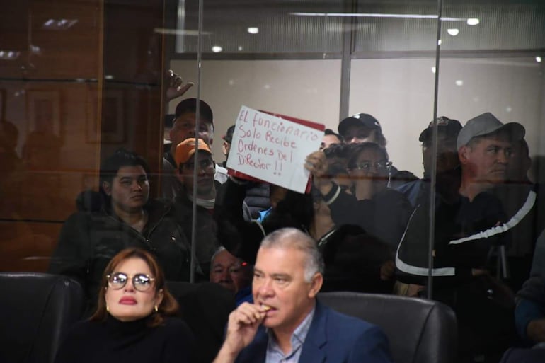 Mujer con gafas grandes y hombre canoso con cigarrillo están sentados, rodeados de personas con cartel de protesta en un ambiente tenso.