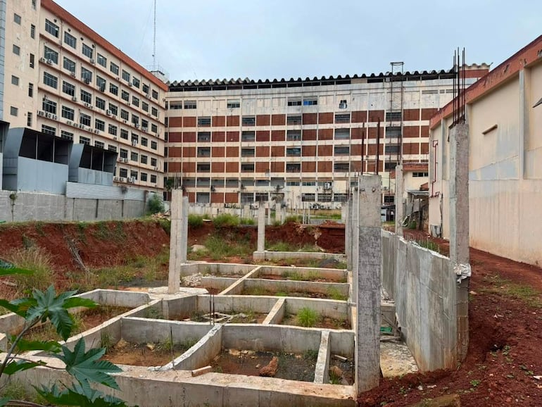 Construcción inconclusa con cimientos de concreto y varillas de hierro, rodeada de vegetación en un entorno urbano.