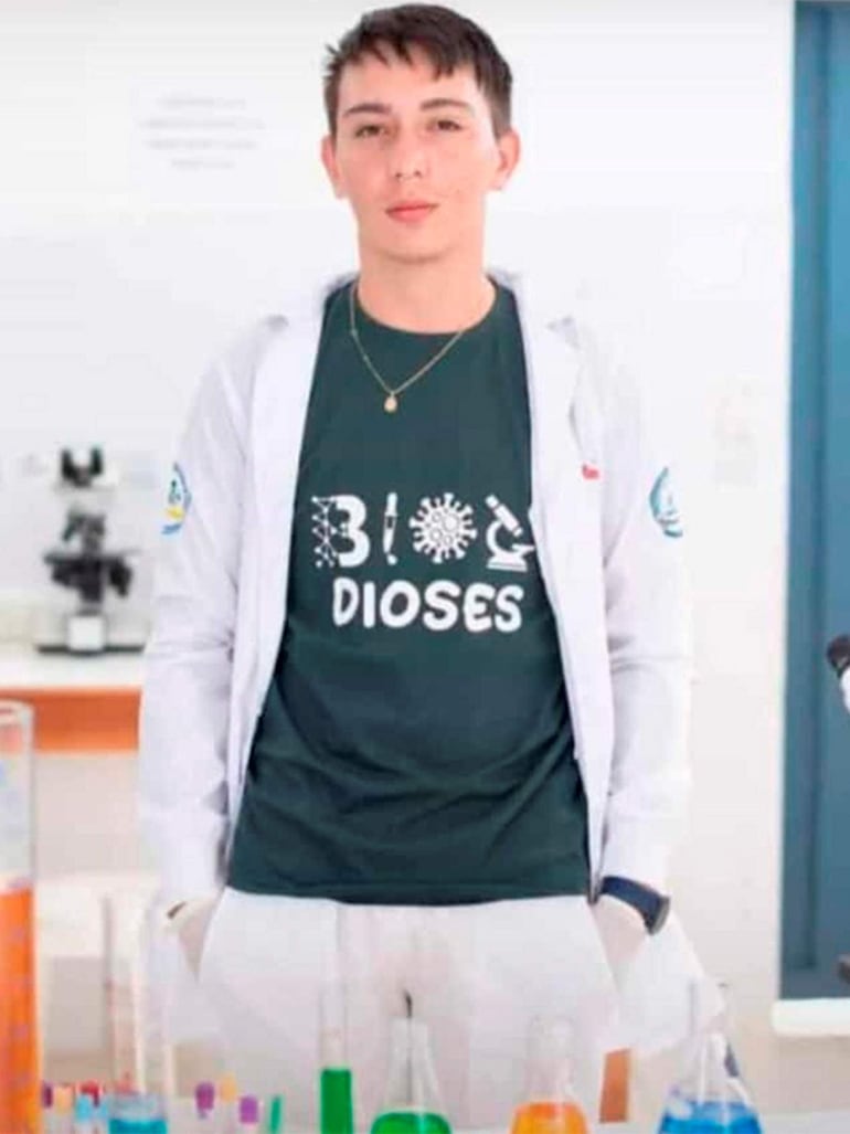 Joven en laboratorio con camiseta verde 'BI DIOSES', lab coat blanco y expresión serena, rodeado de frascos con líquidos de colores.