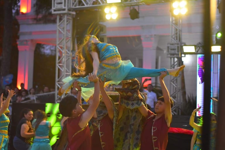 Cuatro jóvenes en el escenario realizan una acrobacia, una mujer vestida de azul es levantada por tres hombres de camisetas marrones.