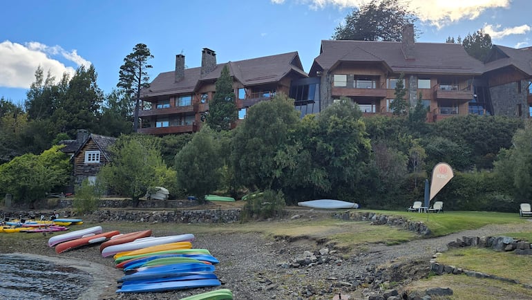Paisaje de playa con kayaks de colores brillantes en la orilla, vegetación alrededor y cabaña de madera al fondo.