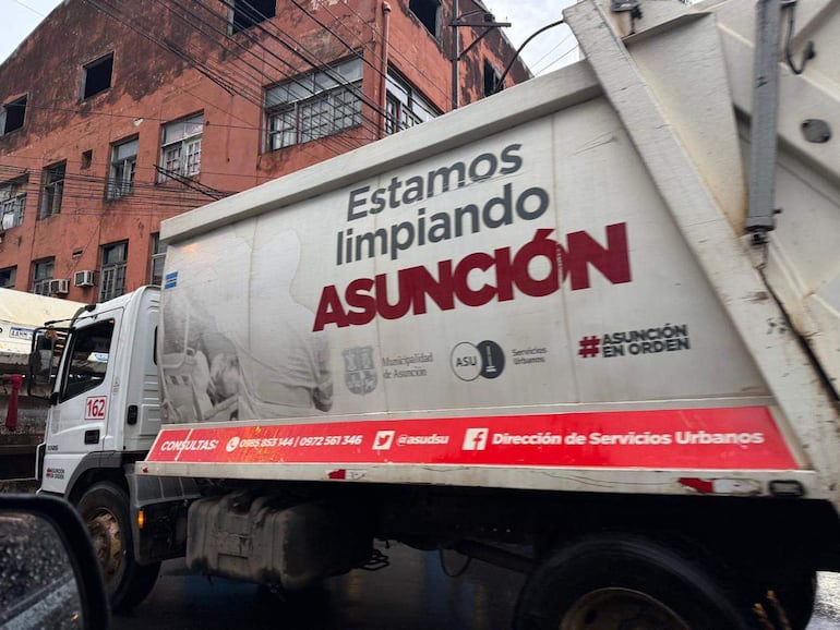 Camión de recolección de basura blanco con slogan 'Estamos limpiando ASUNCIÓN', estacionado en un entorno urbano lluvioso.