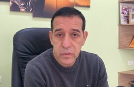 Celso Morales, con suéter gris y expresión seria, sentado en una silla de oficina con fondo verde claro y estantes.