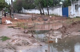 Barricada de malla naranja delimita zona de obras en una calle con pavimento levantado y agua estancada.
