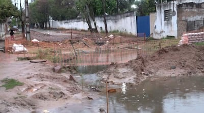Barricada de malla naranja delimita zona de obras en una calle con pavimento levantado y agua estancada.