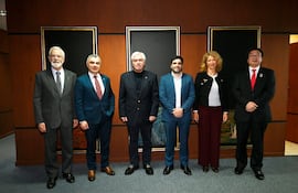 Seis personas vestidas formalmente sonríen en sala decorada con tonos madera y obras abstractas.