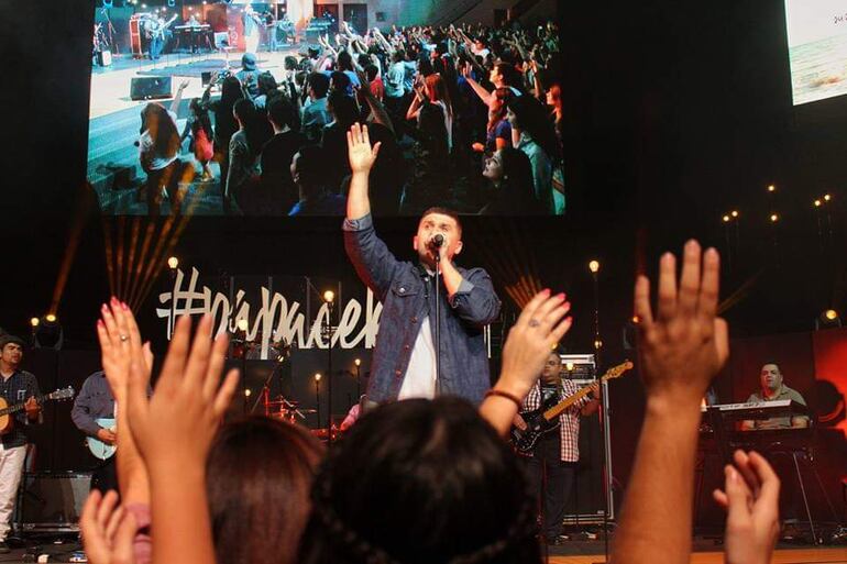 Cantante con micrófono en mano, rodeado de músicos; manos del público se levantan en señal de entusiasmo.