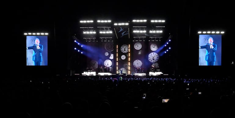 Ricardo Montaner con micrófono en mano, rodeado de músicos, mientras el público graba con teléfonos en un ambiente iluminado.