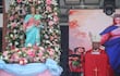 Hombre con atuendo rojo y birrete habla ante altar con la Virgen María y arreglos florales en evento religioso.