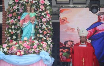 Hombre con atuendo rojo y birrete habla ante altar con la Virgen María y arreglos florales en evento religioso.