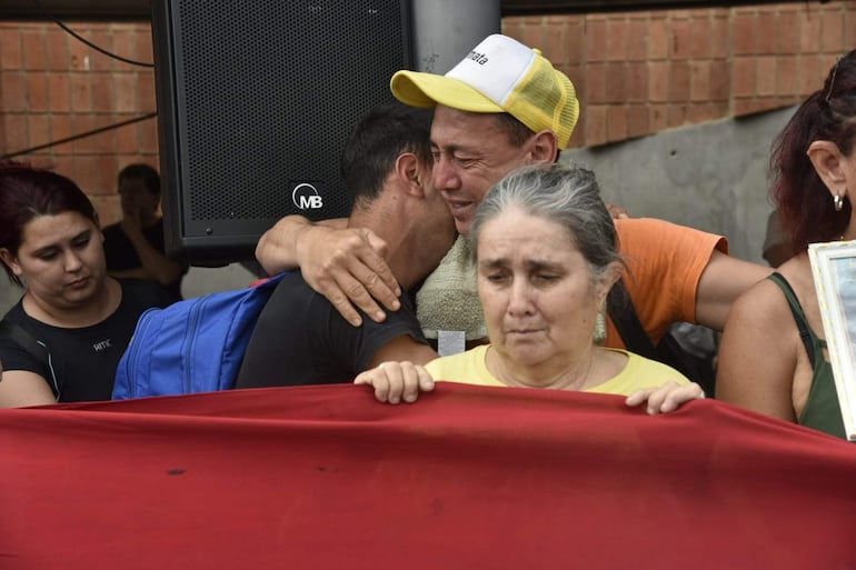Grupo de cinco personas en un acto emocional, con un hombre abrazando a otro y una mujer mayor sosteniendo un cartel.