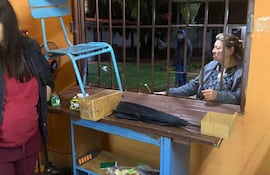 Dos mujeres conversan en un interior de color naranja, una con camiseta roja y chaleco negro, la otra con blusa clara. Un hombre se asoma con chaqueta azul.