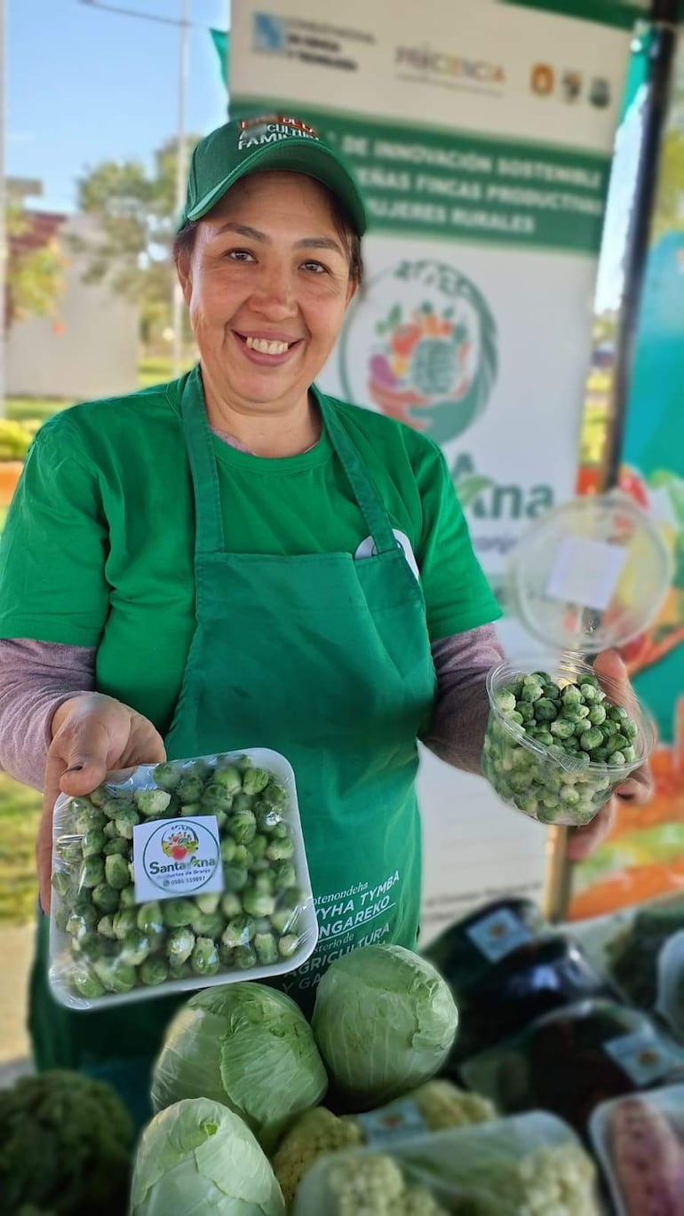 Mujer sonriente con gorra y delantal verde, sostiene recipientes con brócoli en un mercado al aire libre.
