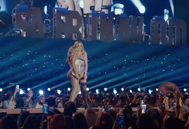 Sabrina Carpenter y Madonna en el escenario, con atuendos llamativos y luces brillantes, ante una gran audiencia que sostiene teléfonos móviles.