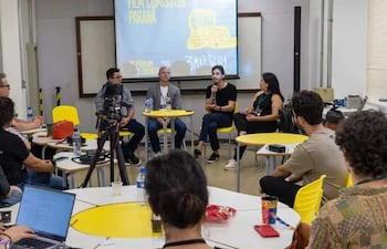 Panel de discusión con cuatro personas, incluidas un hombre hablando con micrófono y una mujer con cabello largo y vestido negro.