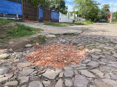 Calle Joel Estigarribia con escombros y cercas azules, en un entorno urbano tranquilo con árboles y edificios de colores.