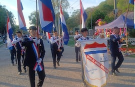 Jóvenes en chaquetas oscuras marchan, sosteniendo banderas paraguayas y un estandarte de Mons. Dr. Ángel Muzzolón en un ambiente colorido.