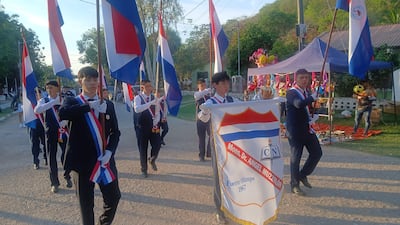 Jóvenes en chaquetas oscuras marchan, sosteniendo banderas paraguayas y un estandarte de Mons. Dr. Ángel Muzzolón en un ambiente colorido.