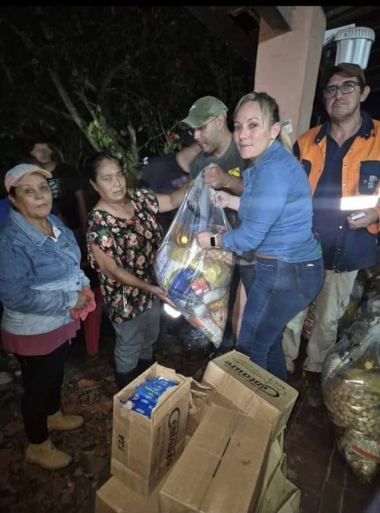 Mujer en el centro entrega alimentos a un grupo de personas en un entorno natural iluminado por luces.