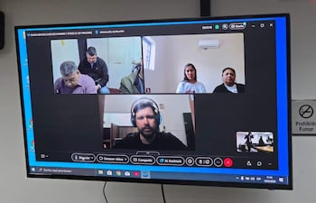 Una persona habla en videoconferencia, rodeada de otros cuatro participantes, todos en un ambiente formal. Cartel 'Prohibido Fumar' visible.