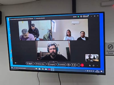 Una persona habla en videoconferencia, rodeada de otros cuatro participantes, todos en un ambiente formal. Cartel 'Prohibido Fumar' visible.