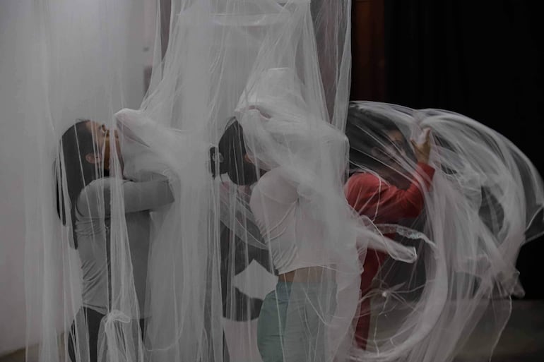 Tres mujeres interactúan con un tejido semitransparente en un ambiente interior, dos visten camisetas claras y una camiseta roja.