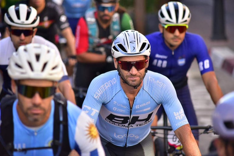 Ciclistas en espera, uno en camiseta azul con logo 'BH COLOMA TEAM' y casco blanco, listos para la carrera en ambiente natural.