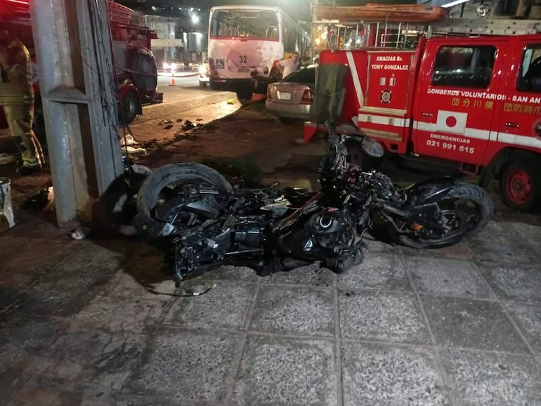 Motocicleta accidentada en la calle, con camión de bomberos y autobús en fondo, iluminado por luces artificiales.