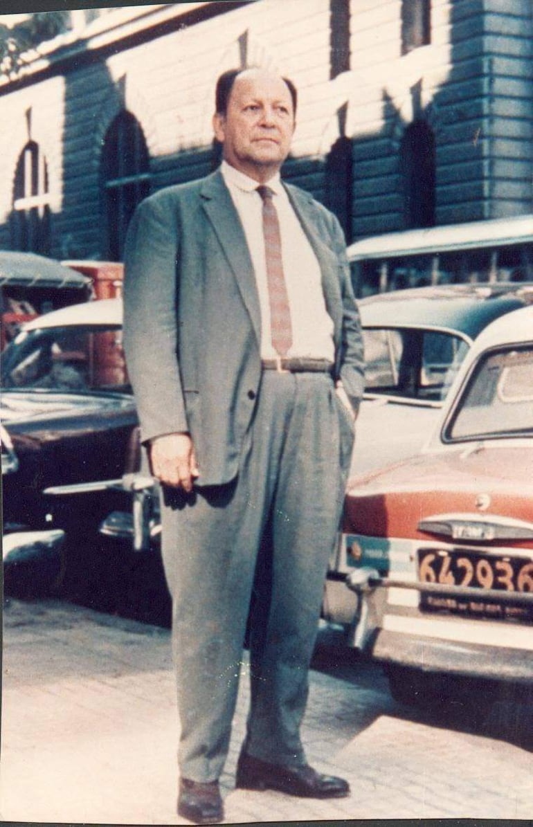 Hombre en traje gris de pie junto a autos antiguos, con una suave sonrisa en un entorno urbano histórico.