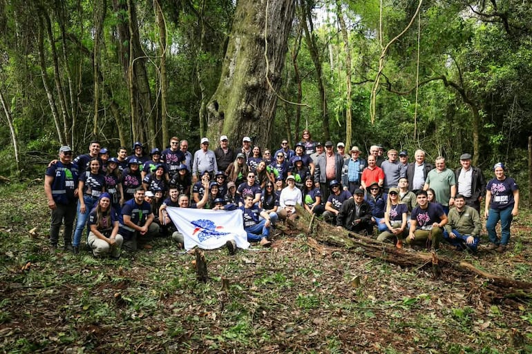 Cincuenta personas en camisetas oscuras, sonriendo, rodeadas de árboles en un bosque, sosteniendo una bandera blanca colorida.