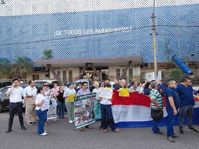 Grupo de personas con uniformes médicos sosteniendo carteles y una bandera de Paraguay, manifestándose junto a un edificio público.