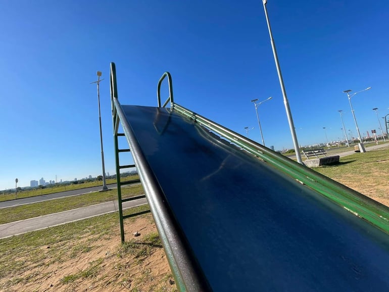 Tobogán de metal verde en área recreativa vacía, reflejando cielo azul sin presencia de personas.