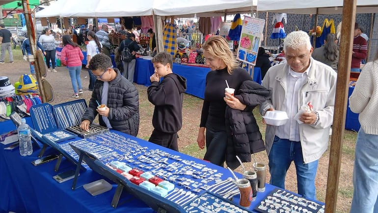 Niño con gafas y mujer de chaqueta negra sostienen objetos en un mercado al aire libre. Hombre mayor observa.