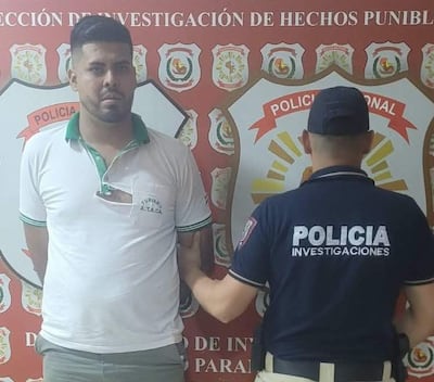 Hombre con camisa blanca y rota bajo custodia policial, mientras un agente de pie supervisa el procedimiento en un ambiente de detención.