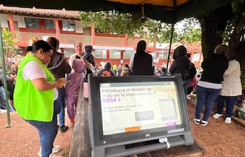 Mujer con chaleco verde fluo y camiseta blanca acompaña a otra mujer que sostiene un niño en un entorno electoral activo.