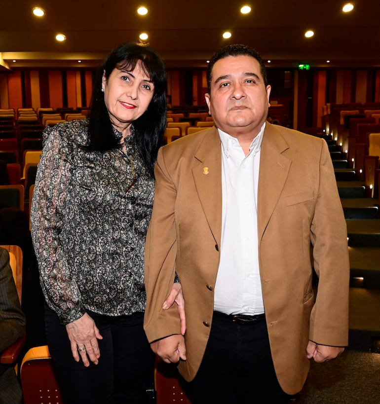 Mujer con blusa de patrón sonriendo junto a hombre en abrigo marrón, ambos en un evento con asientos y luz tenue.