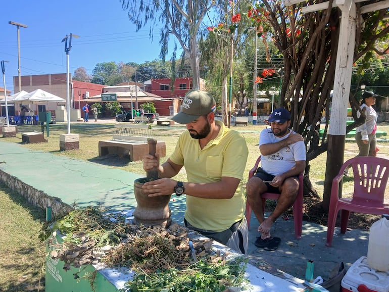 Hombre con sombrero y camiseta amarilla usando un mortero, mientras otro hombre vestido de blanco lo observa, rodeados de vegetación y asientos.