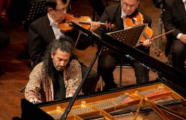 Nima Sarkechik, con chaqueta negra y abrigo colorido, interpreta en piano de cola negro, mientras músicos lo acompañan formalmente detrás.