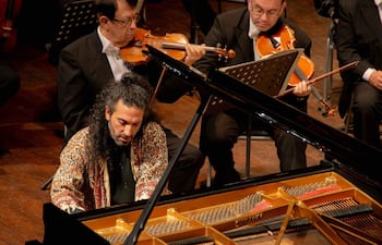 Nima Sarkechik, con chaqueta negra y abrigo colorido, interpreta en piano de cola negro, mientras músicos lo acompañan formalmente detrás.