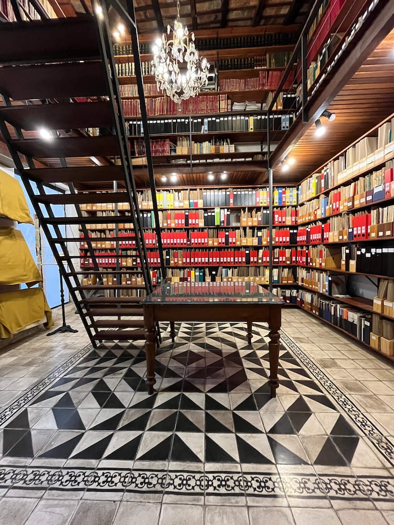 Interior de biblioteca acogedora con estanterías llenas de libros, lámpara de cristal y escalera metálica en un entorno histórico.