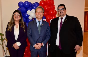Iván Yueh-Jung Lee sonriente en traje azul claro, con mujer de chaqueta oscura y hombre de traje negro con corbata, rodeados de globos coloridos.
