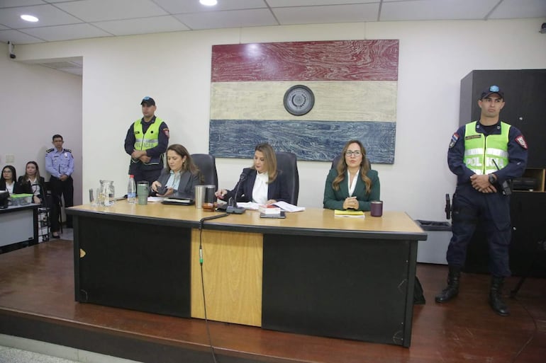 Yolanda Morel y Ana Rodríguez en traje formal discuten documentos mientras un policía observa en un recinto judicial.